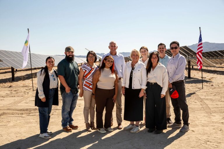 PCE staff gather together while standing in front of solar panels on a bright, sunny day