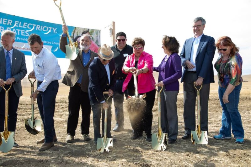 A group of people are smiling as they dig shovels into the dirt with a large celebratory banner in the background