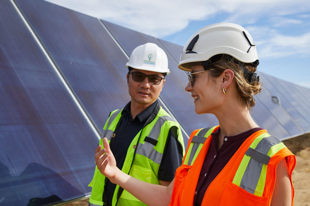 Two Peninsula Clean Energy employees in safety vests and helmets conducting fieldwork at a solar energy facility.