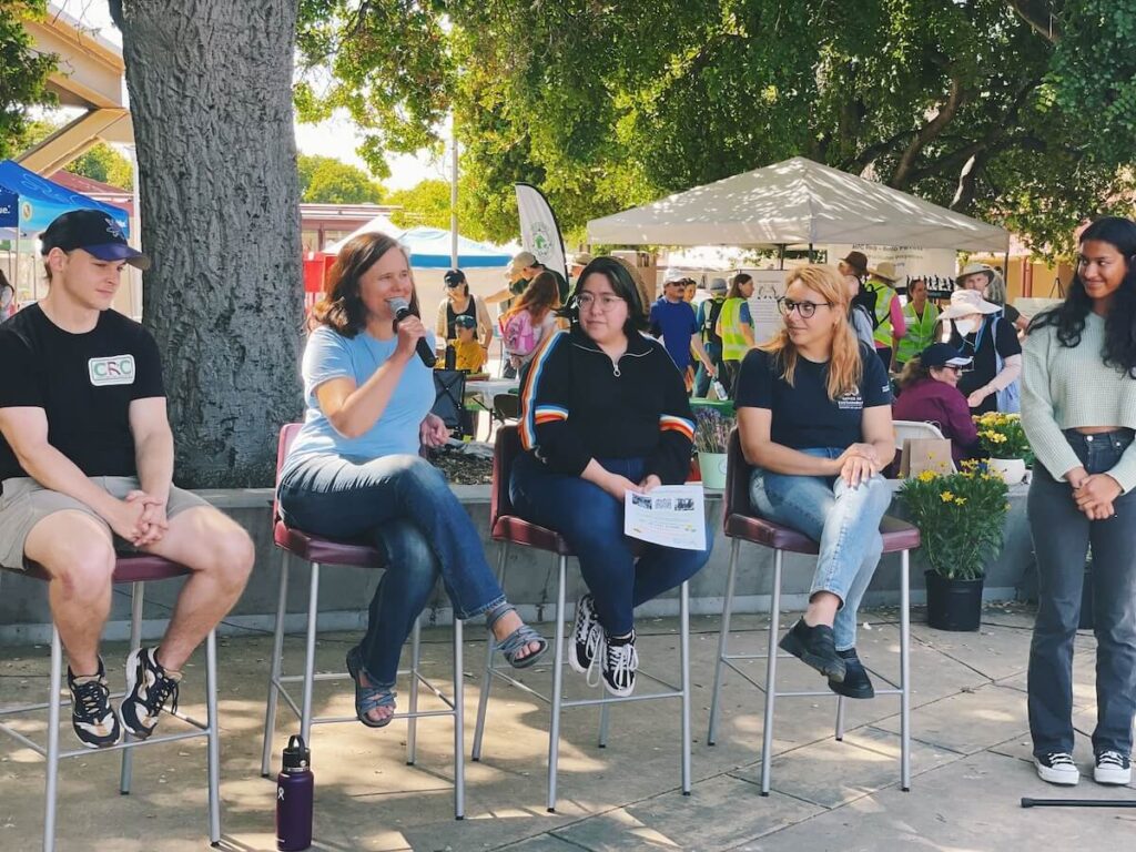 Four panelists sit in tall chairs at an outdoor community event, and a woman in the center holds a microphone to her mouth as she answers a question