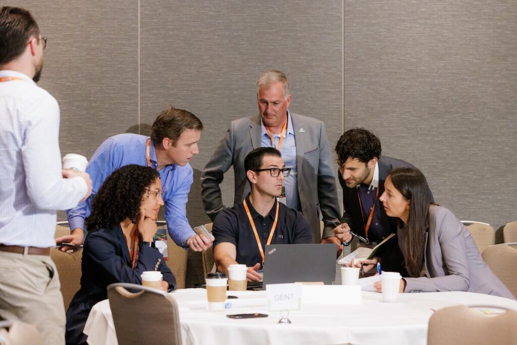 A group of professionals wearing lanyards sit at a table and discuss something displayed on a laptop screen