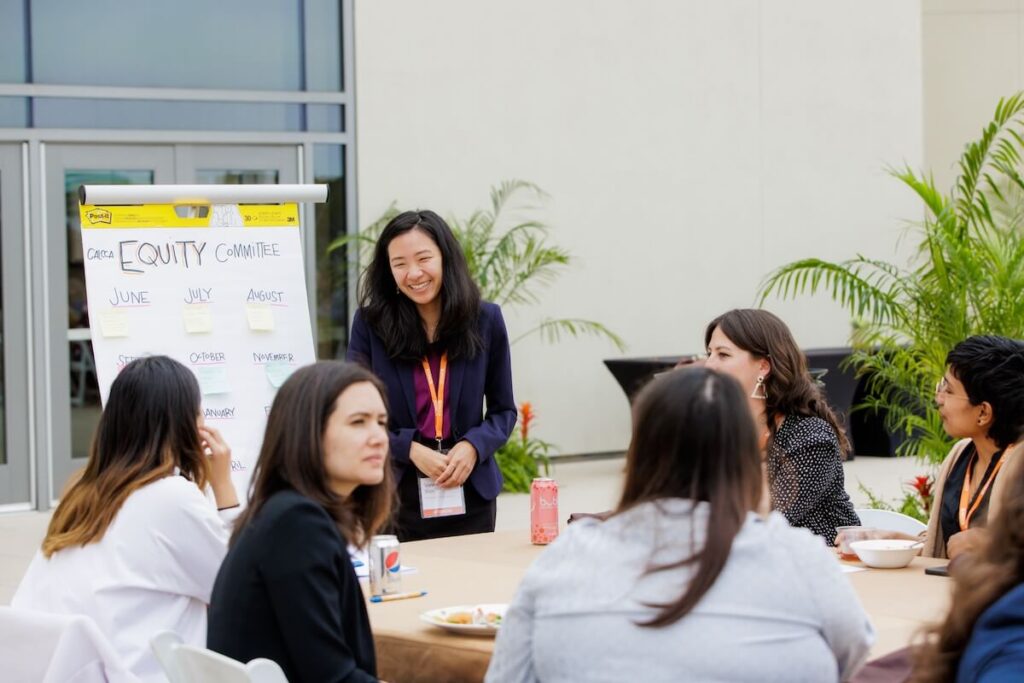 A group of women are seated around a table and female facilitator stands near a board that reads "Equity Committee"