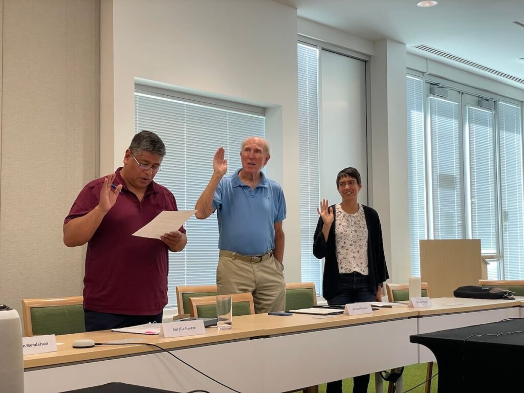 Two adult men and an adult woman stand at a long table while raising their hands, seemingly voting while at a meeting