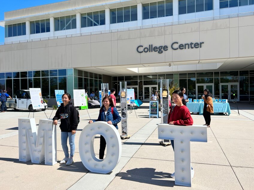 Three female students stand behind oversized statues of letters that read "M-O-T" in front of a large building labeled "College Center."