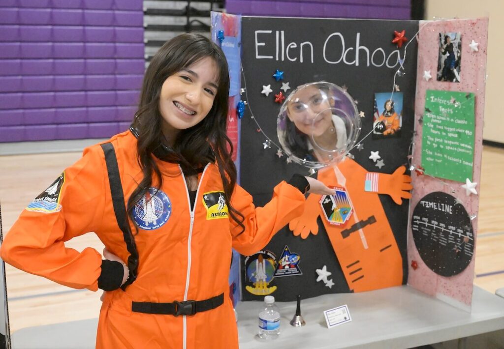 A middle school aged girl wears an orange space jumpsuit and gestures towards her science project board that reads Ellen Ochoa